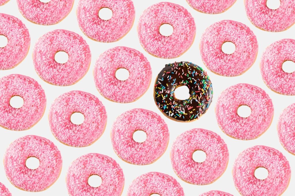 crowd of pink donuts with one chocolate donut amongst them