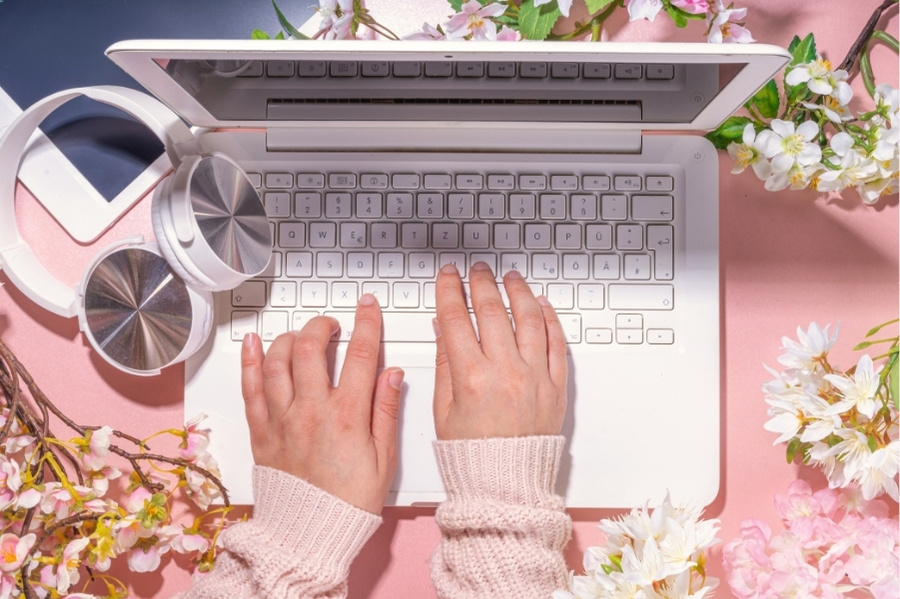 woman's hands typing on a white laptop surrounded by blossom on a pink desk